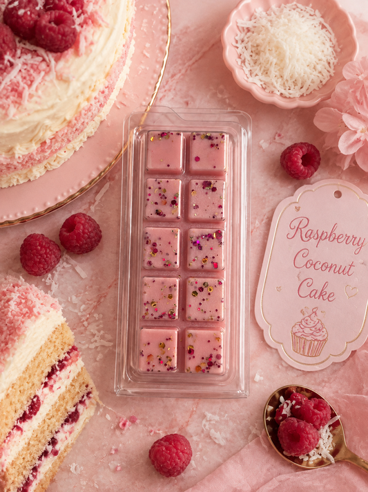 A clear plastic clamshell package containing pink raspberry coconut cake wax melt bar styled on a pink surface with raspberries, cake slices, shredded coconut, and a printed tag reading 'Raspberry Coconut Cake'.