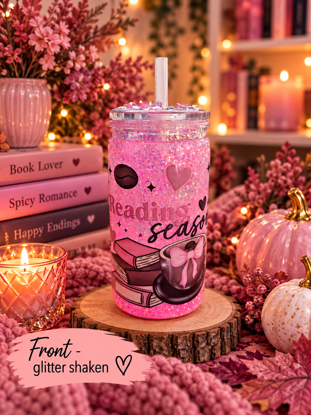 A pink glitter-shaken tumbler cup with a straw, decorated with a coffee cup and books graphic and the words 'reading season,' displayed on a wooden slice in a cozy fall-themed setting with candles, pumpkins, and books.