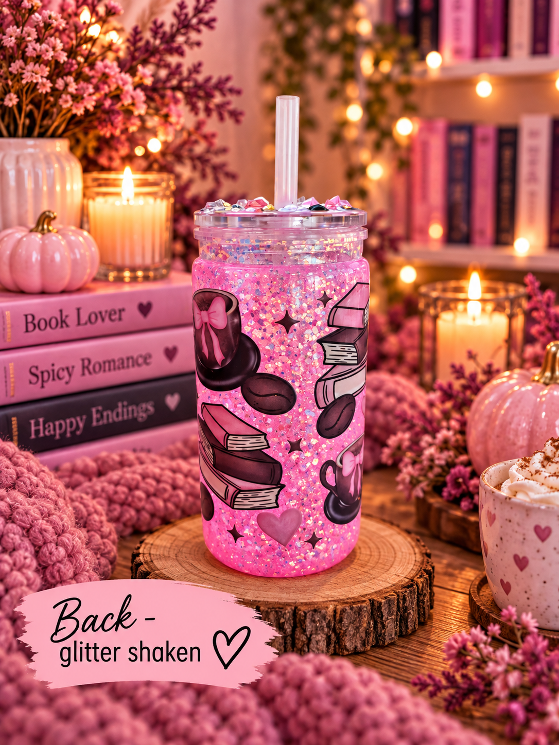 A pink glitter-filled tumbler with book-themed graphics sits on a wooden slice in a cozy, warmly lit setting with candles, books, pumpkins, and floral decor. A pink banner at the bottom reads 'Back - glitter shaken'.