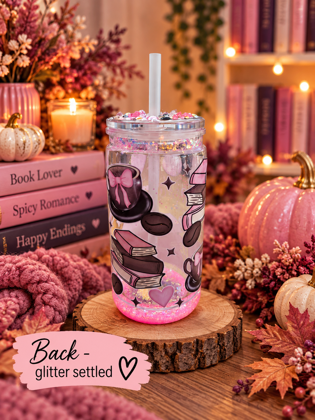 A clear glitter tumbler with a white straw, decorated with pink and brown book illustrations and glitter, set on a wooden slice in a cozy autumn-themed setting with pumpkins, candles, and books.