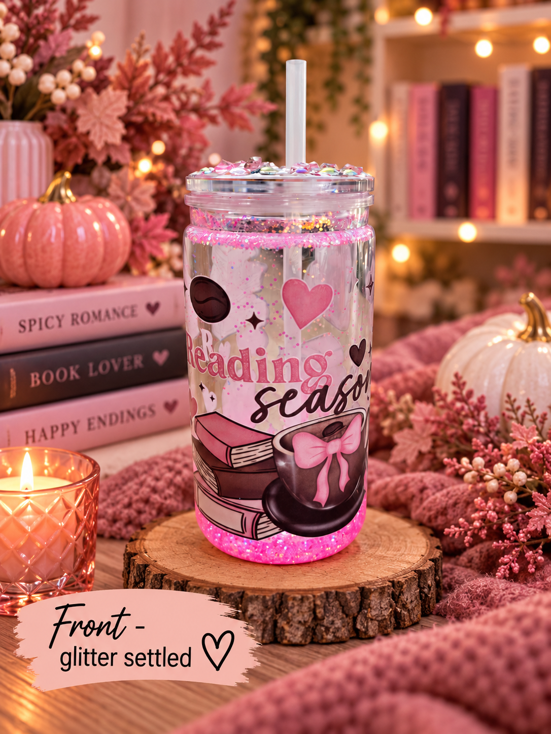 A clear tumbler with a white straw and glitter-filled rim, printed with a cozy autumn reading-themed design featuring pink hearts, books, a coffee cup with a bow, and the words 'reading season.' The tumbler sits on a wood slice in a styled fall scene with pumpkins, books, a candle, and warm string lights in the background.
