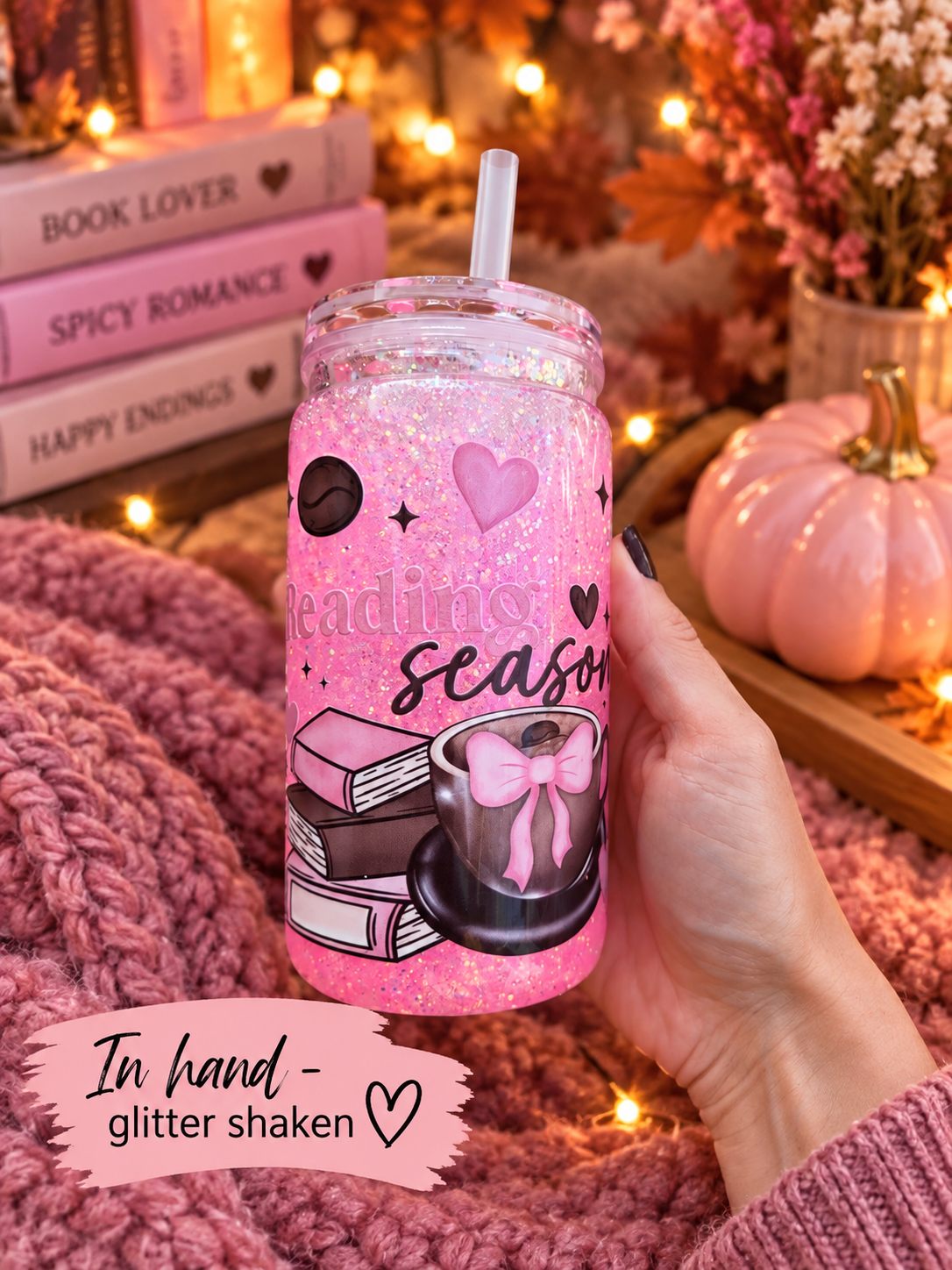 A hand holds a pink glitter tumbler with a straw, decorated with illustrations of books, a coffee cup with a bow, and the words 'reading season'. The background is a cozy autumn-themed setting with warm fairy lights, a pink knit blanket, a small pumpkin, and books.