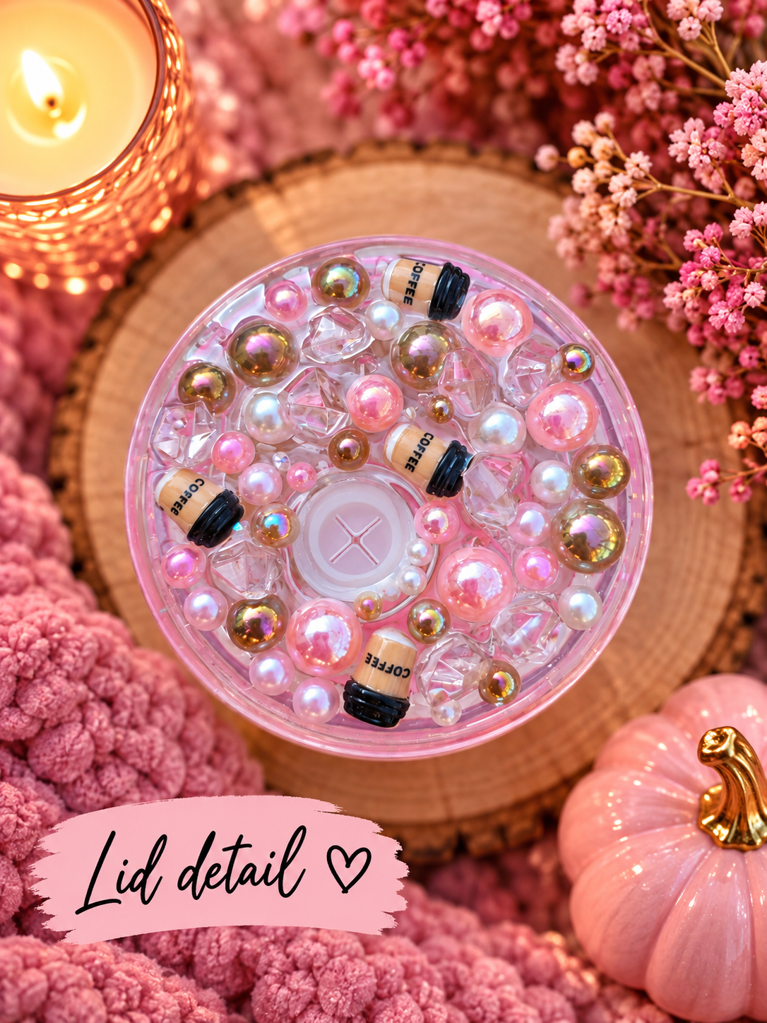 A close-up of a round clear plastic jar lid filled with pink and iridescent beads, pearl-like spheres, and small black-and-gold capsule charms, styled on a wood slice with pink flowers, a candle, and a pink pumpkin. Text overlay reads 'Lid detail ♥'.