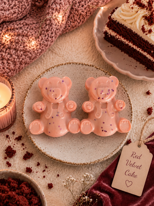 Two teddy bear-shaped wax melts decorated with purple glitter, presented on a speckled ceramic plate. The setting is styled as a cozy dessert table with a slice of red velvet cake, a candle, a knit blanket, and a tag reading 'Red Velvet Cake'.