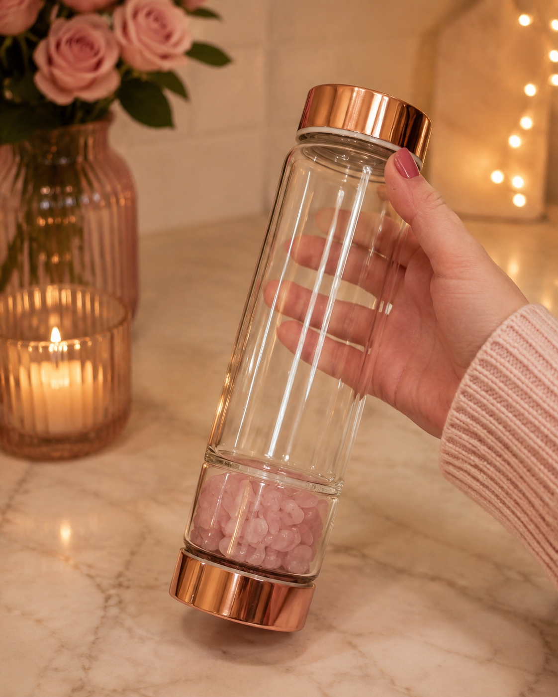 A hand holds a clear glass water bottle with rose gold metal caps and pink salt crystals at the bottom, set on a marble countertop with a warm, cozy background featuring a lit candle and flowers.