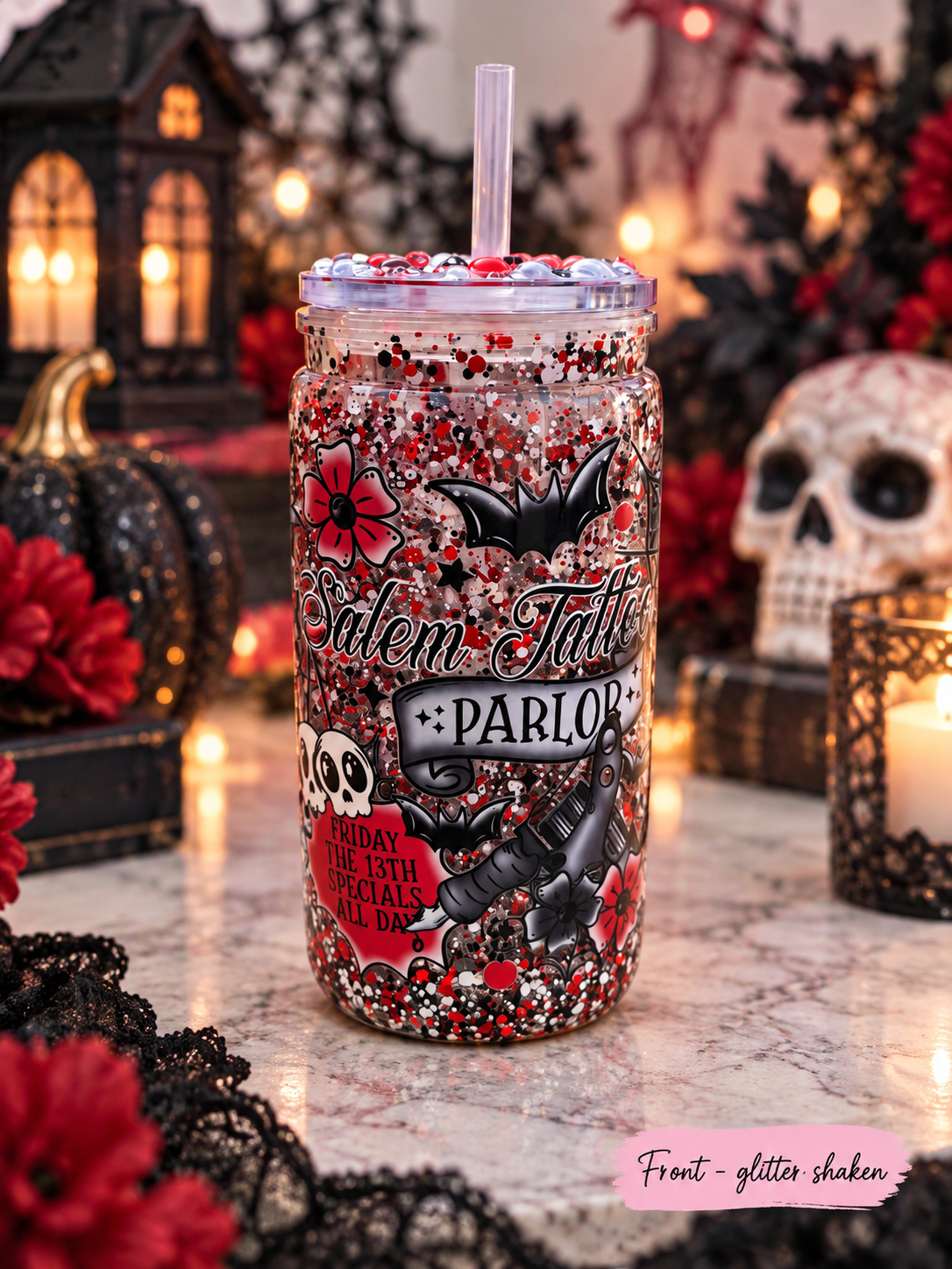 A Halloween-themed glitter shaker mason jar tumbler with a clear lid and straw, decorated with red, black, and white glitter, skulls, bats, and the text 'Salem Tattoo Parlor'. The tumbler is set on a marble surface surrounded by seasonal decorations including a skull, candles, and pumpkins.