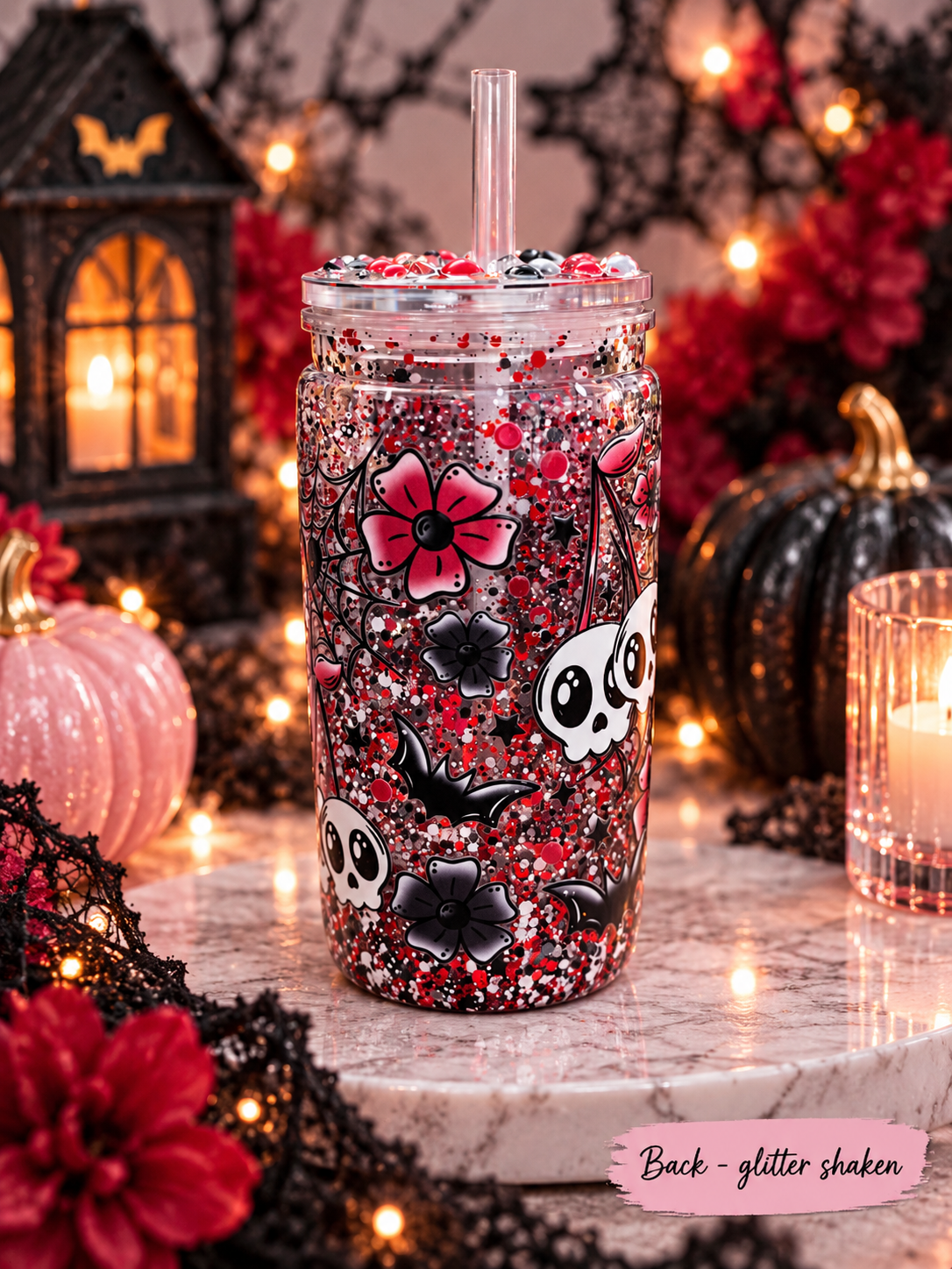 A clear glitter tumbler with a straw, decorated with red and black glitter, skulls, bats, and pink flowers, set on a marble surface with a Halloween background.