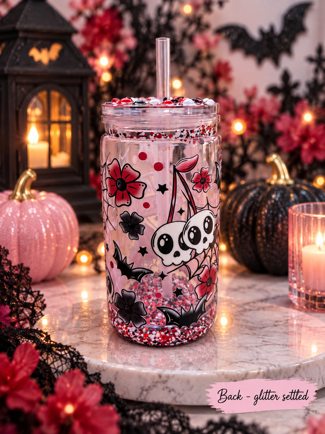A clear glitter-set tumbler with a straw, decorated with skulls, flowers, and bats on a pink background, displayed on a marble surface with Halloween decorations in the background.