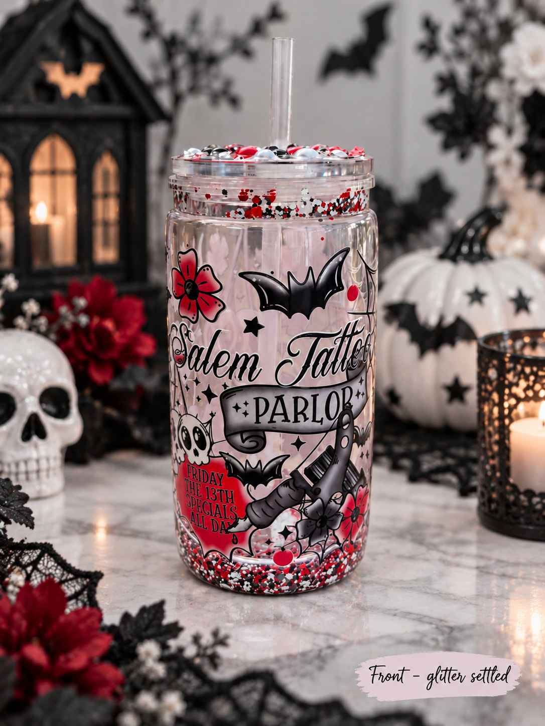 A clear Halloween-themed mason jar tumbler with a straw, featuring gothic illustrations of Salem Tattle Parlor, black bats, red roses, and glitter-filled base and rim. The tumbler is set on a marble surface surrounded by Halloween decorations including a skull, lantern, pumpkins, and black lace.