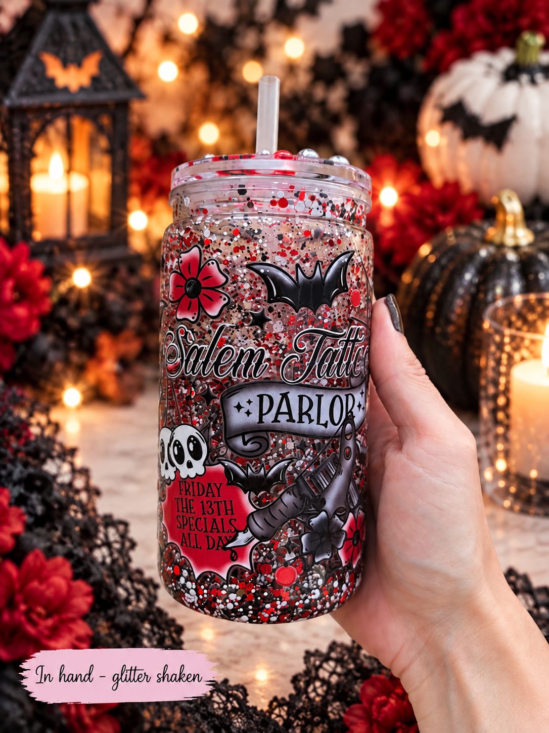 A hand holds a clear glitter tumbler with a straw, decorated with Halloween-themed graphics including bats, skulls, roses, and the text 'Salem Tattoo Parlor' and '10th Day of the Month Specials All Day.' The background is a warm, festive Halloween scene with lanterns, candles, lace, pumpkins, and red flowers.