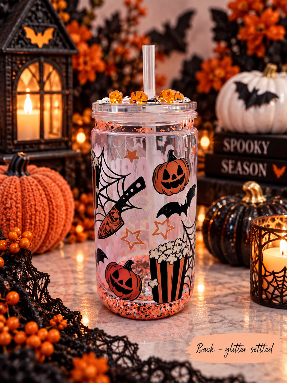 A Halloween-themed clear plastic tumbler with a clear straw, decorated with orange jack-o'-lanterns, black bats, spiderwebs, and a popcorn bucket graphic. The tumbler is filled with glitter and sits on a marble surface surrounded by festive Halloween decorations, including pumpkins, a lantern, and candles.