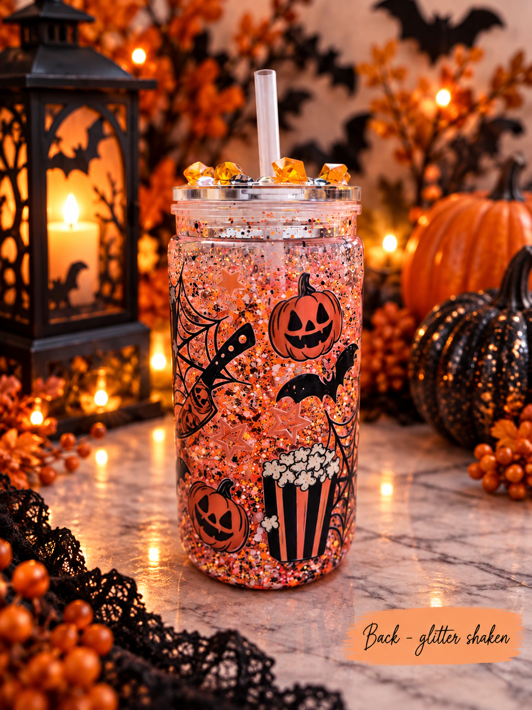 A Halloween-themed glitter shaker tumbler cup with a clear straw, decorated with black jack-o'-lanterns, bats, and popcorn graphics over a pink glitter base. The cup is styled on a marble surface with a black lantern, pumpkins, and warm lights in the background.