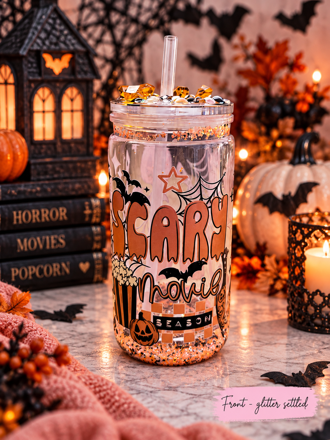 A clear glitter-filled mason jar tumbler with a straw, decorated with 'Scary Movie Season' text, bats, and popcorn graphics, set in a cozy Halloween-themed environment with pumpkins, candles, and books.