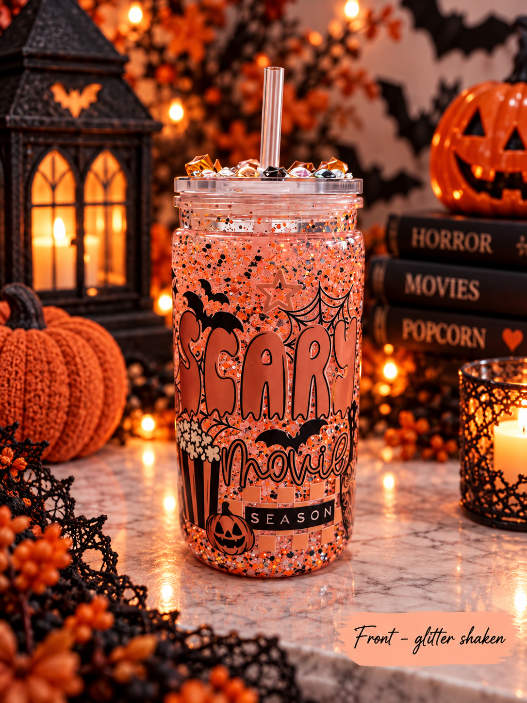 A clear plastic tumbler with a straw, filled with orange glitter and decorated with Halloween-themed graphics including bats, spiders, popcorn, and the words 'SCARY movies SEASON'. The tumbler is styled on a marble surface with a black lantern, pumpkins, books, and a candle in a spooky autumn setting.