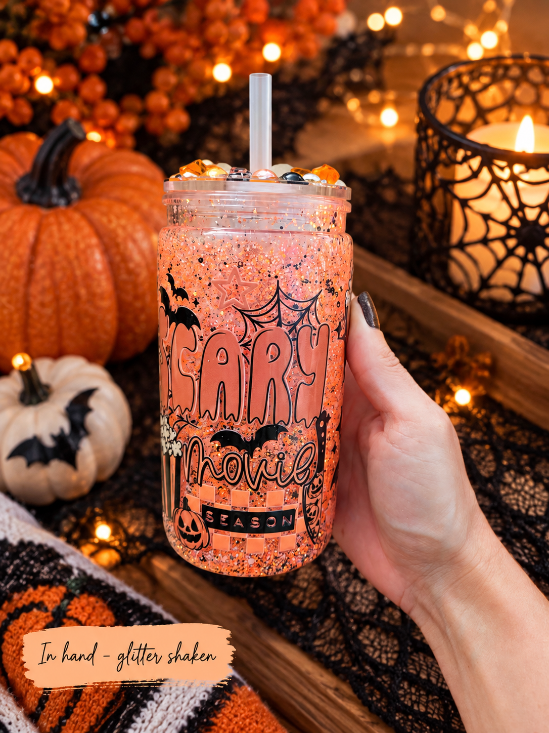 A hand holds a clear glitter-tinted mason jar tumbler with a straw, decorated with orange and black Halloween graphics including bats and the text 'Halloween Season.' The background is a styled autumn scene with pumpkins, string lights, a candle, and black lace, with an overlay note reading 'In hand - glitter shaken.