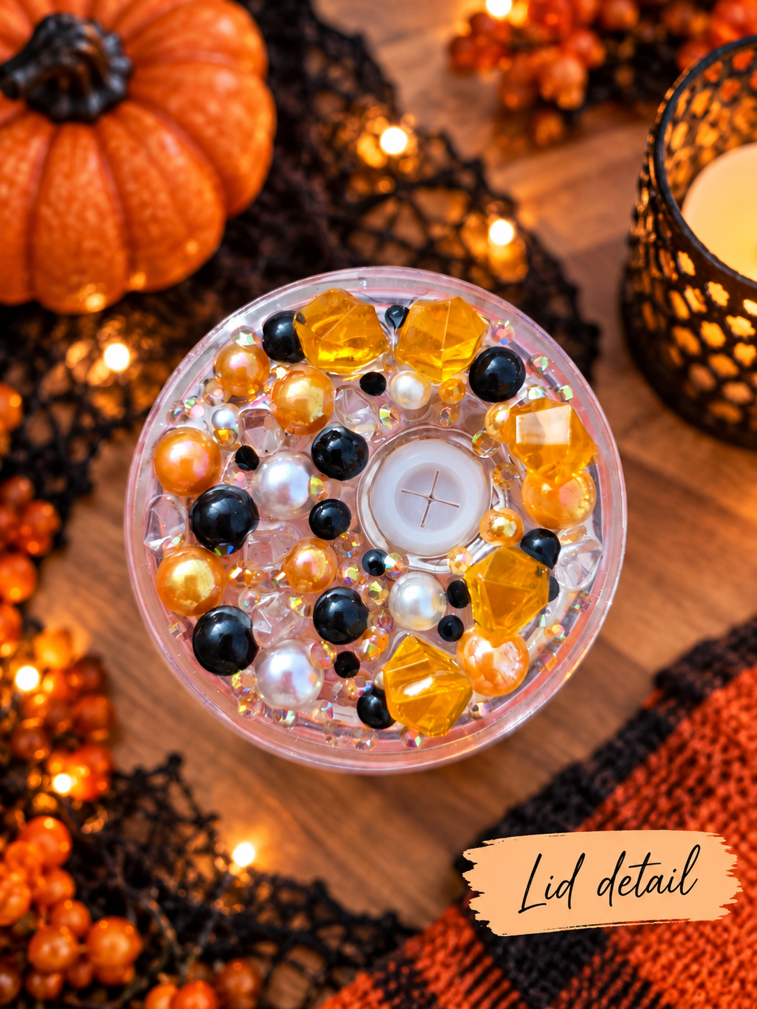 A top-down close-up of a decorative tumbler lid with embedded amber faceted crystals, black and white pearls, and a small white tea light in the center, surrounded by a warm autumn-themed styled scene with a pumpkin, lace, string lights, and a candle holder. A label in the corner reads 'Lid detail'.