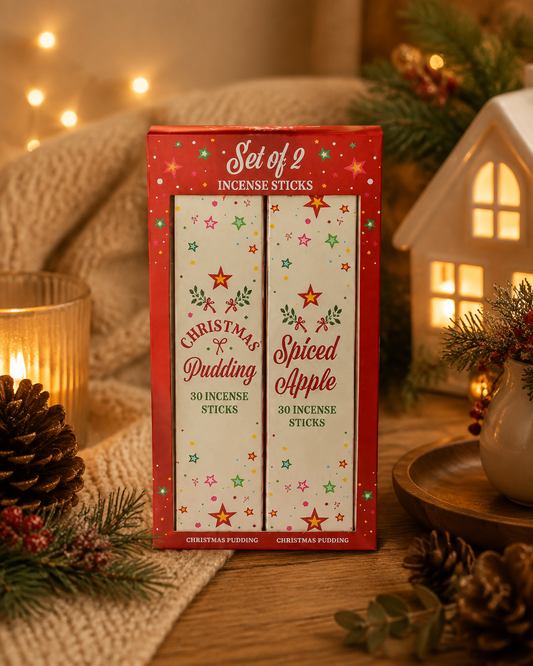 A boxed set of two Christmas-themed incense sticks stands on a wooden surface with holiday decor around it, including pine branches, a lit candle, a small house-shaped lantern, and warm string lights in the background.