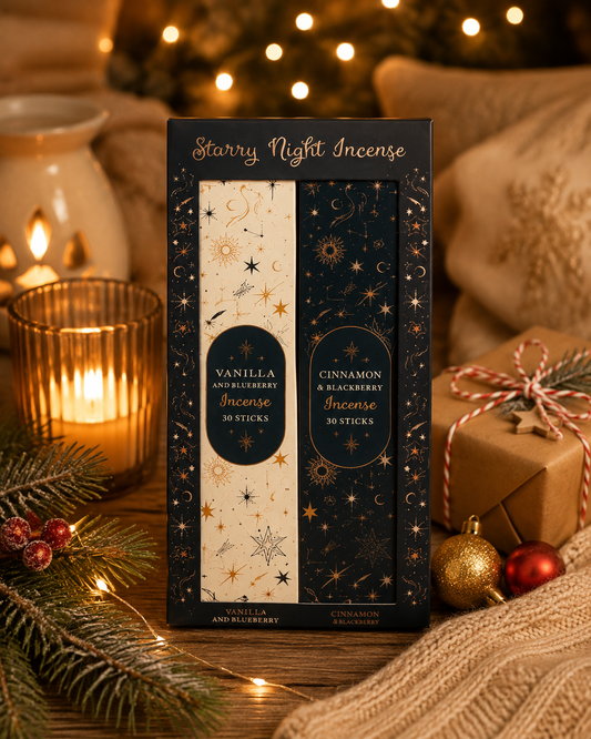 A boxed set of two scented incense sticks stands on a table surrounded by holiday decor, including a lit candle, pine branches, ornaments, and a wrapped gift. The box reads 'Starry Night Incense' and shows vanilla and cinnamon and blackberry incense.