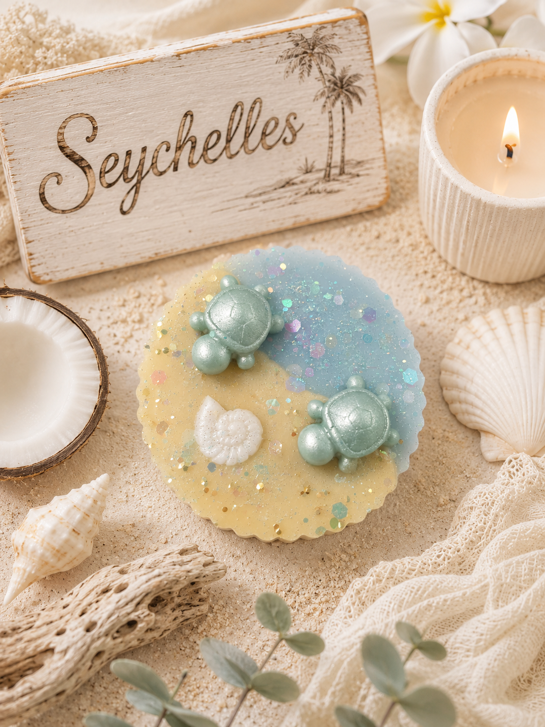 A round handmade wax melt with a yin-yang design in pastel blue and pale yellow, decorated with three mint-green sea turtle figures and glitter, styled on a sandy surface with seashells, a wooden 'Seychelles' sign, and a lit candle.