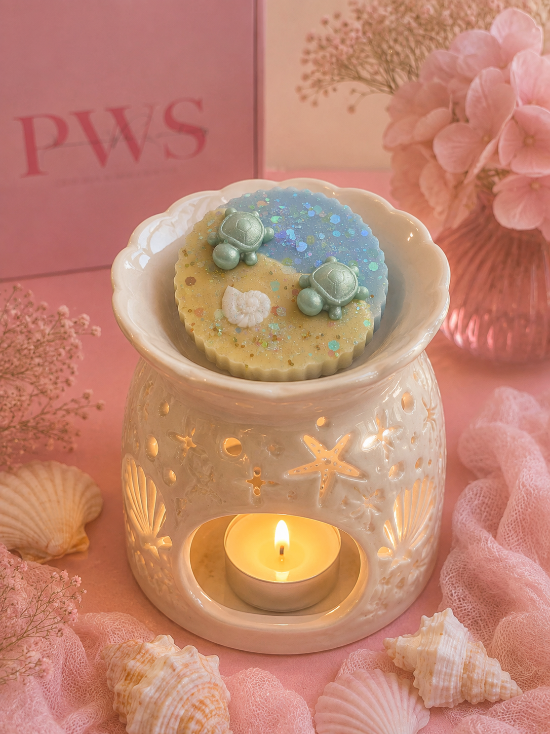 A ceramic wax warmer with starfish and shell cutouts, lit with a small tealight candle inside, holding a round decorative wax melt topped with three sea turtle figures. The scene is styled with pink fabric, seashells, dried flowers, and a pink box in the background.