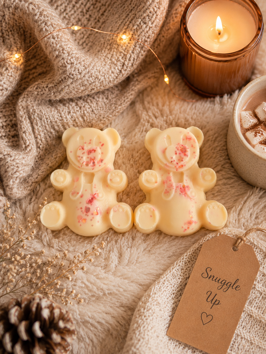 Two teddy bear-shaped wax melts in pale cream with pink sprinkles, styled on a cozy beige knit blanket with a lit candle, a cup of hot chocolate, fairy lights, and a 'Snuggle Up' gift tag.