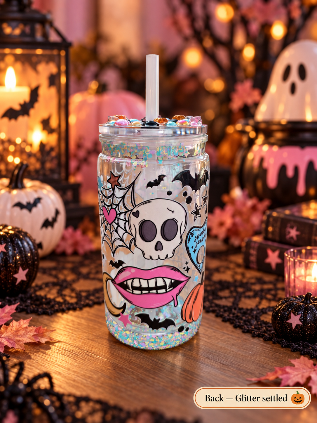 A Halloween-themed glitter tumbler cup with a white straw, decorated with a skull, spiderwebs, bats, pink lips, and a blue pumpkin, set on a wooden table surrounded by pumpkins, black lace, and warm holiday lights.