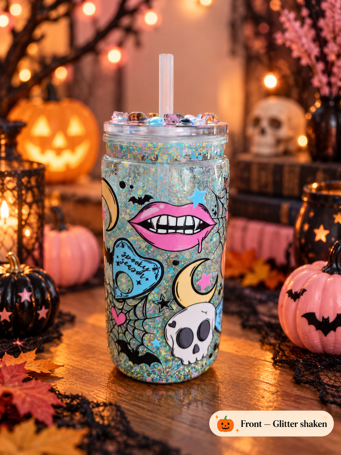 A Halloween-themed glitter tumbler with a clear lid and straw, decorated with pink lips, a skull, crescent moons, bats, and a blue heart. The tumbler is set on a wooden table surrounded by pumpkins, candles, and autumn decorations.