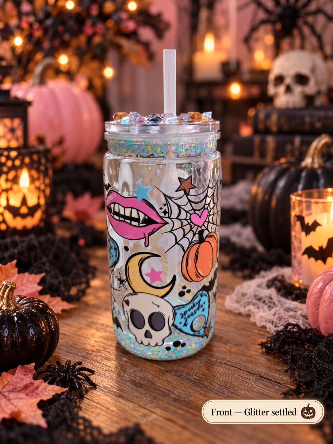 A clear glitter tumbler with a white straw, decorated with Halloween-themed illustrations including a skull, moon, spiderweb, lips, and an orange pumpkin, set on a wooden table surrounded by pumpkins, candles, and black lace in a cozy autumn setting.