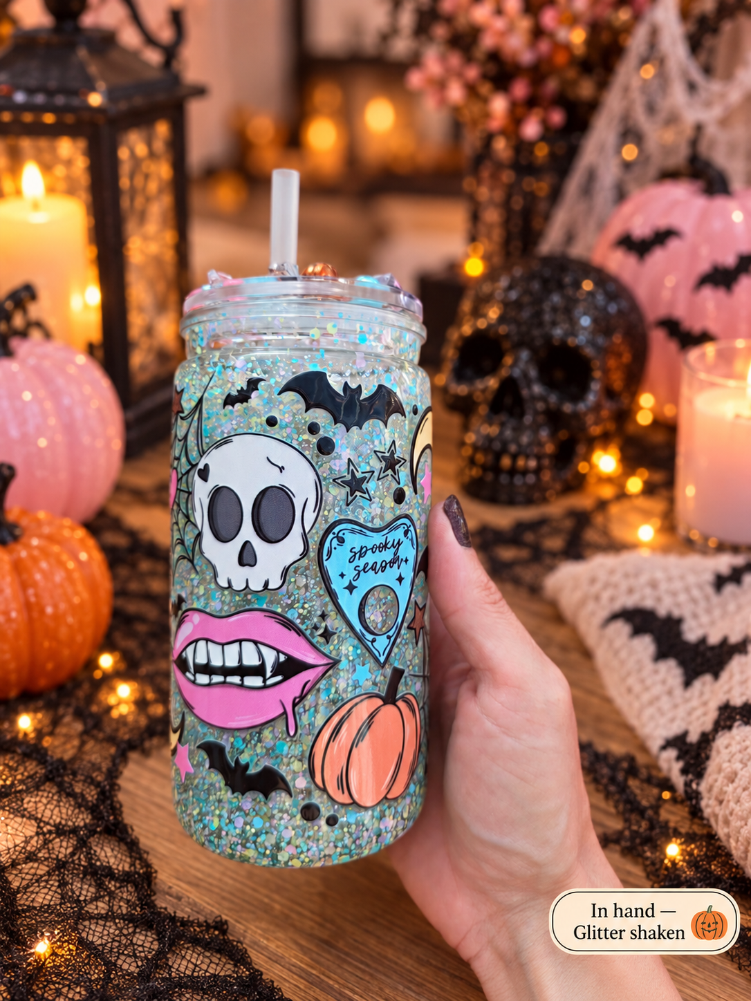 A hand holds a glittery clear tumbler with a straw decorated with Halloween graphics including a white skull, pink lips, a small orange pumpkin, black bats, and a blue heart that reads 'Spooky Sweetheart'. The background is a cozy, warmly lit Halloween table setting with candles, lanterns, and pumpkins.