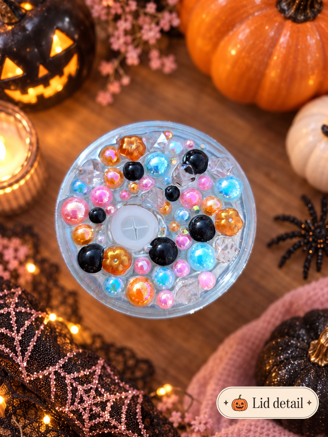 A round clear lid filled with colourful craft beads and sequins, photographed from above on a wooden surface surrounded by Halloween decorations including pumpkins, a candle, spiderweb fabric, and a small 'Lid detail +' label in the corner.
