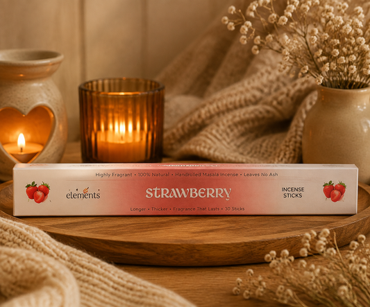 A box of Elements Strawberry Incense Sticks sits on a wooden tray surrounded by cozy home decor, including a lit candle, a amber glass holder, and dried flowers.