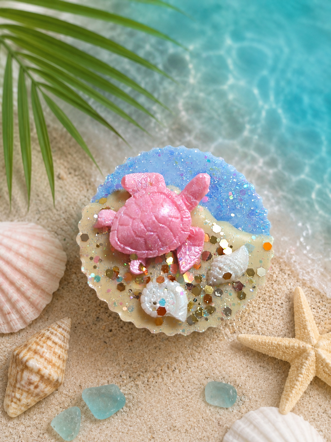 A round beach-themed wax melt sits on sand with shells, a starfish, and turquoise water in the background. The product has a blue glitter top half, a beige bottom half, a pink sea turtle decoration, and white shell accents.