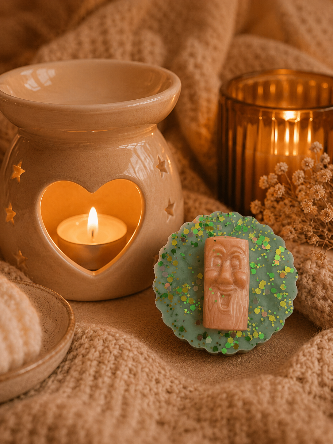 An oval green wax melt with a carved wooden face on a glittery green backing, styled beside a lit beige ceramic candle warmer and a amber glass candle holder on a cozy beige knit blanket.