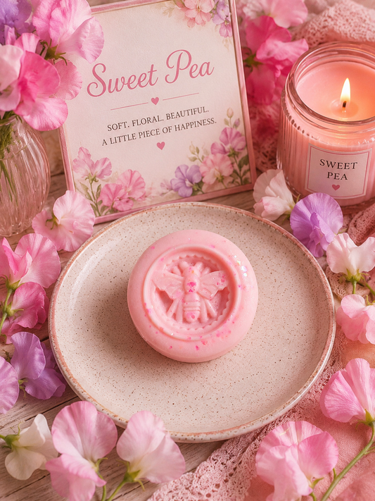 A pink round wax melt with a bee embossing sits on a speckled beige ceramic plate, surrounded by pink flowers, a lit pink candle labeled 'Sweet Pea,' and a matching floral sign.