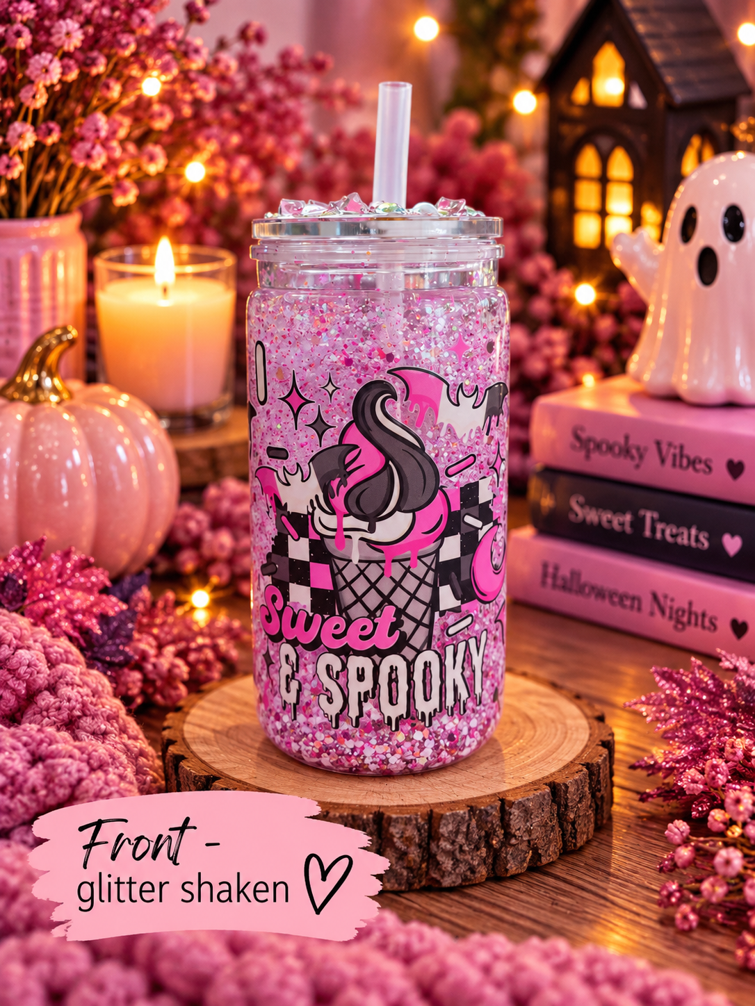 A clear glass mason jar tumbler filled with pink glitter and a clear straw, decorated with a black and pink cartoon character and the words 'Sweet & Spooky.' It sits on a wood slice in a cozy Halloween-themed setting with candles, books, flowers, and a ghost figurine.