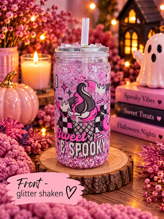 A clear glass mason jar tumbler filled with pink glitter and a clear straw, decorated with a black and pink cartoon character and the words 'Sweet & Spooky.' It sits on a wood slice in a cozy Halloween-themed setting with candles, books, flowers, and a ghost figurine.