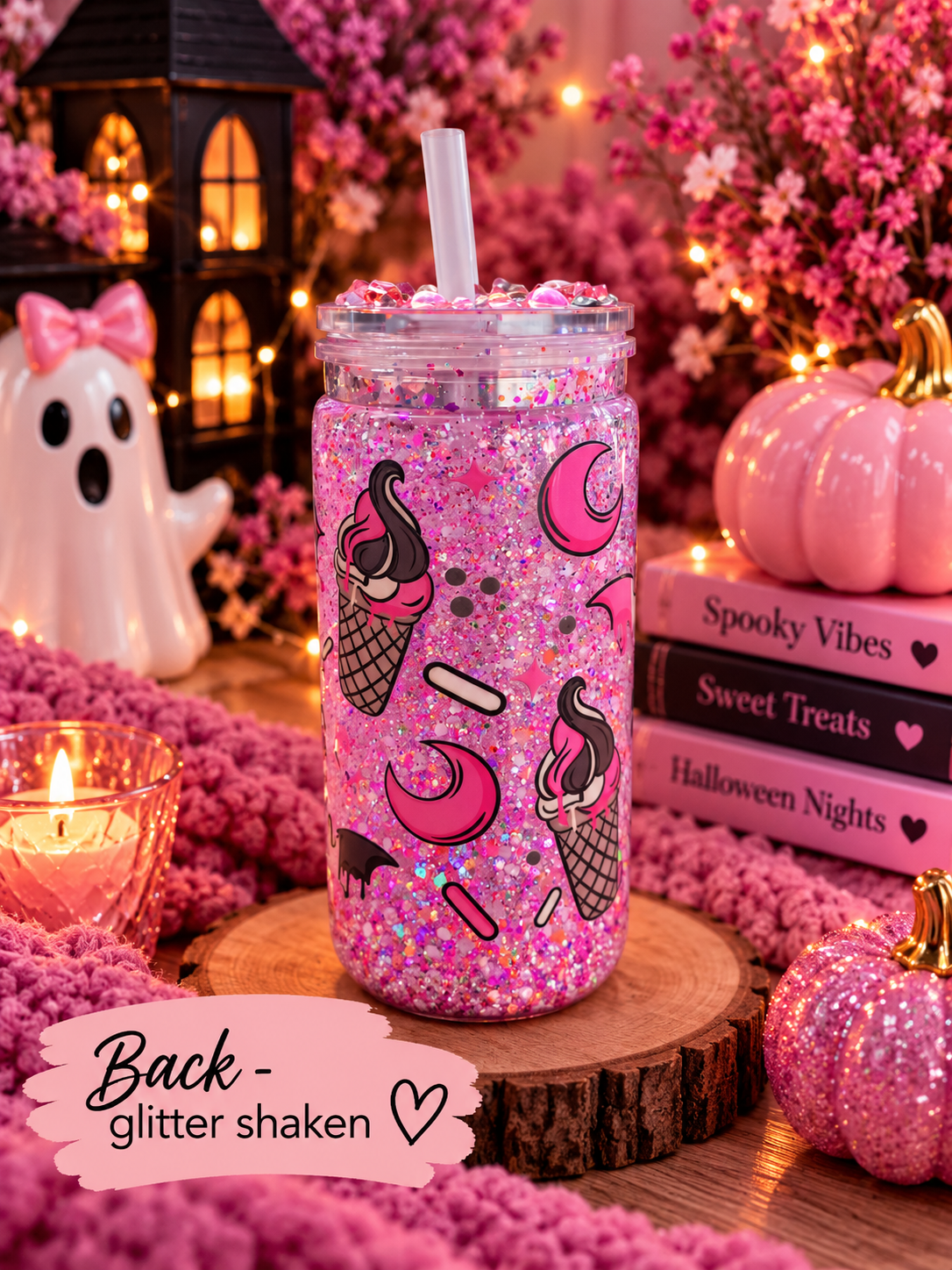 A pink glitter shaker tumbler with ice cream cone and moon illustrations, filled with pink glitter and topped with a clear straw, displayed on a wooden slice in a cozy Halloween-themed setting with books, a ghost lantern, and pink decorations.
