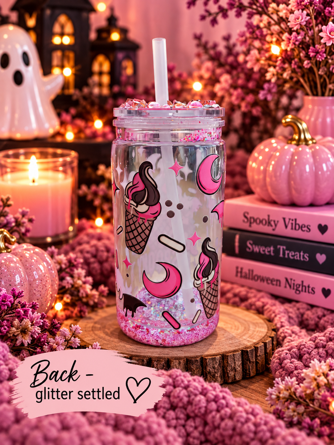 A clear glitter tumbler with a straw, decorated with pink and black ice cream cone and moon illustrations, set on a wooden slice surrounded by pink flowers, candles, and Halloween-themed decor.