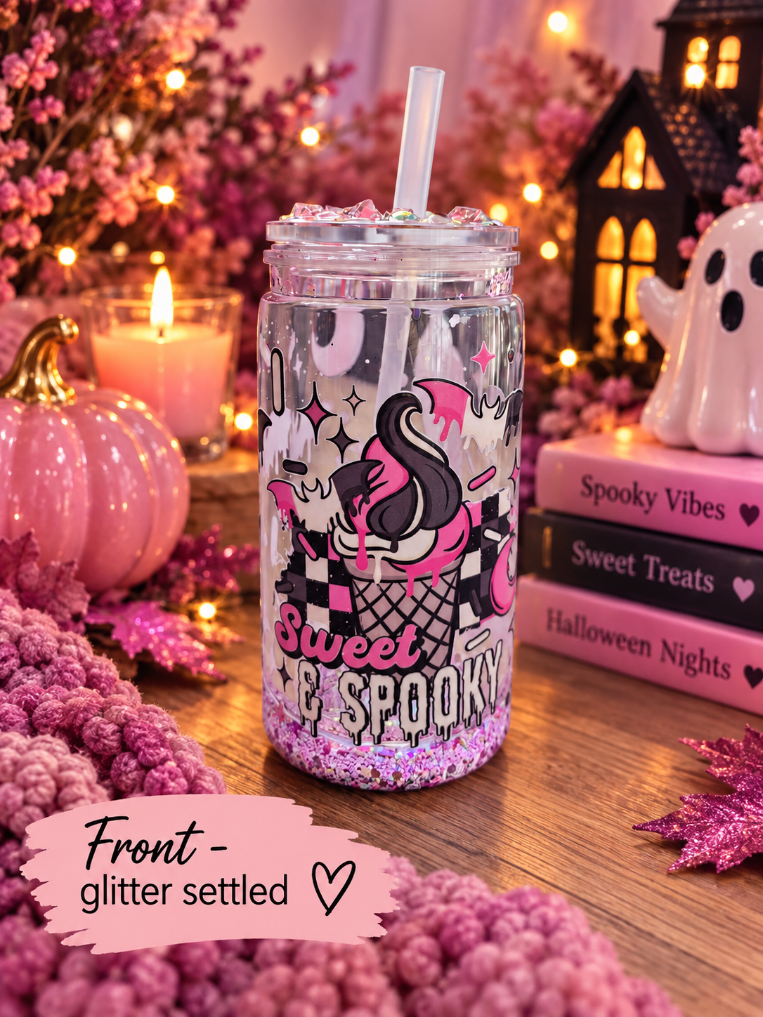 A clear glass mason jar tumbler with a straw, decorated with a pink flamingo, black and pink checkerboard pattern, and the words 'Sweet and Spooky' in glitter, set on a wooden table surrounded by Halloween decorations including pink pumpkins, books, and a ghost figurine.