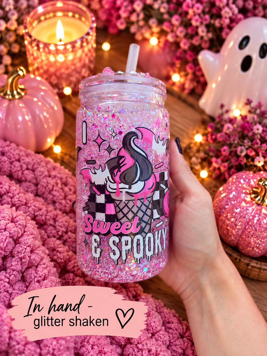 A hand holds a clear glitter tumbler with a straw, decorated with a pink and black cartoon unicorn and the words 'Sweet & Spooky.' The scene is styled in fall colors with pink pumpkins, flowers, candles, and a ghost decoration.