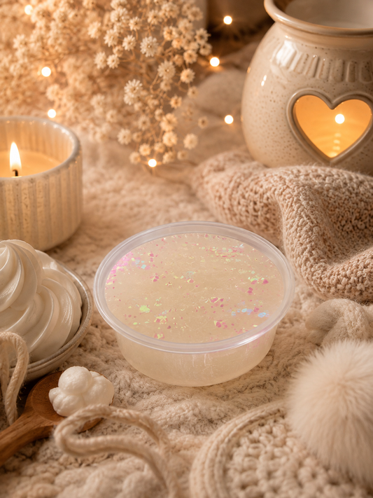A clear plastic tub filled with gel wax melt with small iridescent glitter, styled on a cozy knit blanket with a candle, dried flowers, a ceramic holder, and warm string lights.
