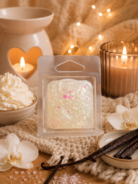 A packaged gel wax melt cube sits on a cozy knit blanket with warm candles, whipped cream, vanilla beans, and white orchids around it.