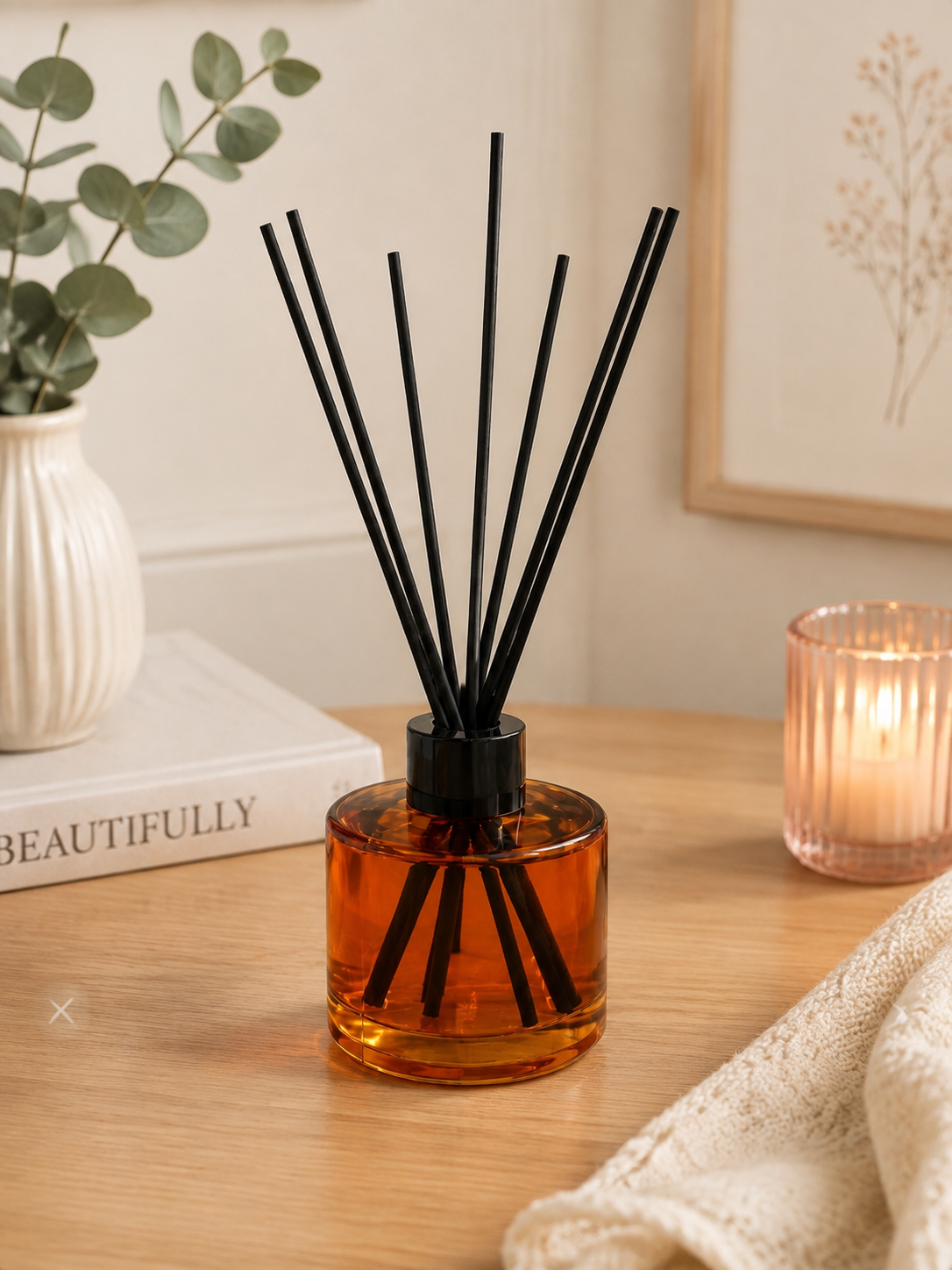 Amber glass reed diffuser bottle with black diffuser sticks on a wooden table, styled with a white vase of greenery, a book, and a lit pink candle in the background.