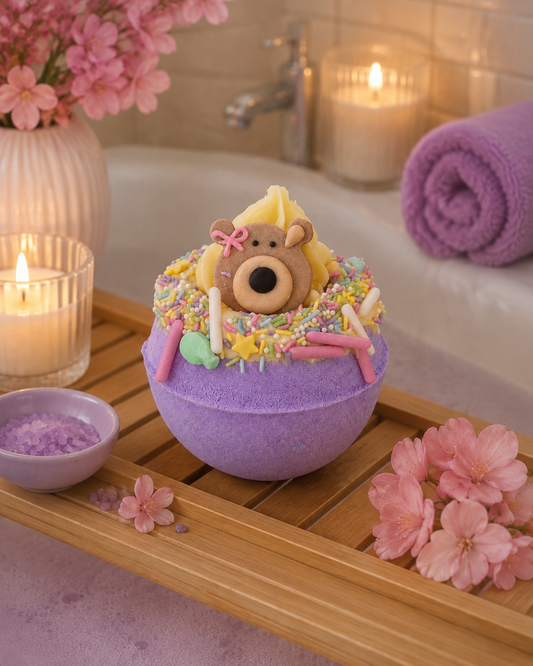 A purple bath bomb shaped like a cupcake sits on a wooden tray in a spa-like setting. The top is decorated with yellow frosting, colorful sprinkles, a small teddy bear cookie, and pink candy accents.