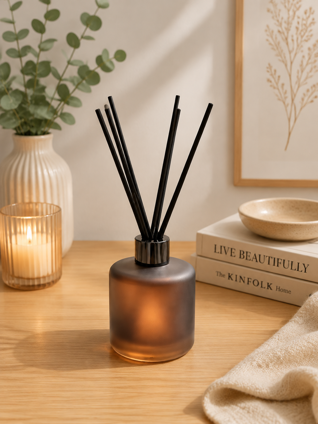 A frosted amber glass reed diffuser with black reeds sits on a wooden table in a styled interior scene with a lit candle, vase, framed botanical print, books, and a folded cloth.