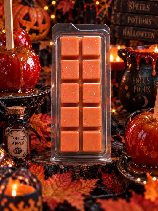 An orange toffee apple scented wax melt bar in a clear plastic clamshell package, displayed on a rustic Halloween-themed table with candy apples, autumn leaves, and themed props.