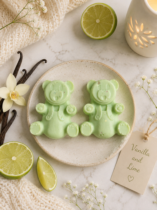 Two pale green bear-shaped wax melts displayed on a round beige plate, surrounded by decorative props including lime slices, vanilla beans, a cream knit fabric, small white flowers, and a tag reading 'Vanilla and Lime'.
