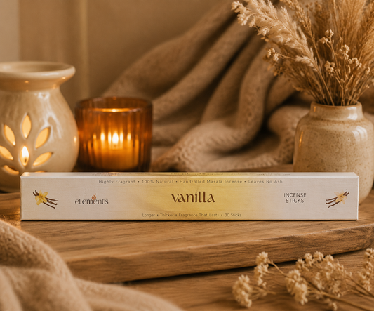 A box of vanilla scented incense sticks from the brand Elements, styled on a wooden surface with a lit amber candle, a cream ceramic holder, and dried grass in a vase, creating a warm, cozy atmosphere.
