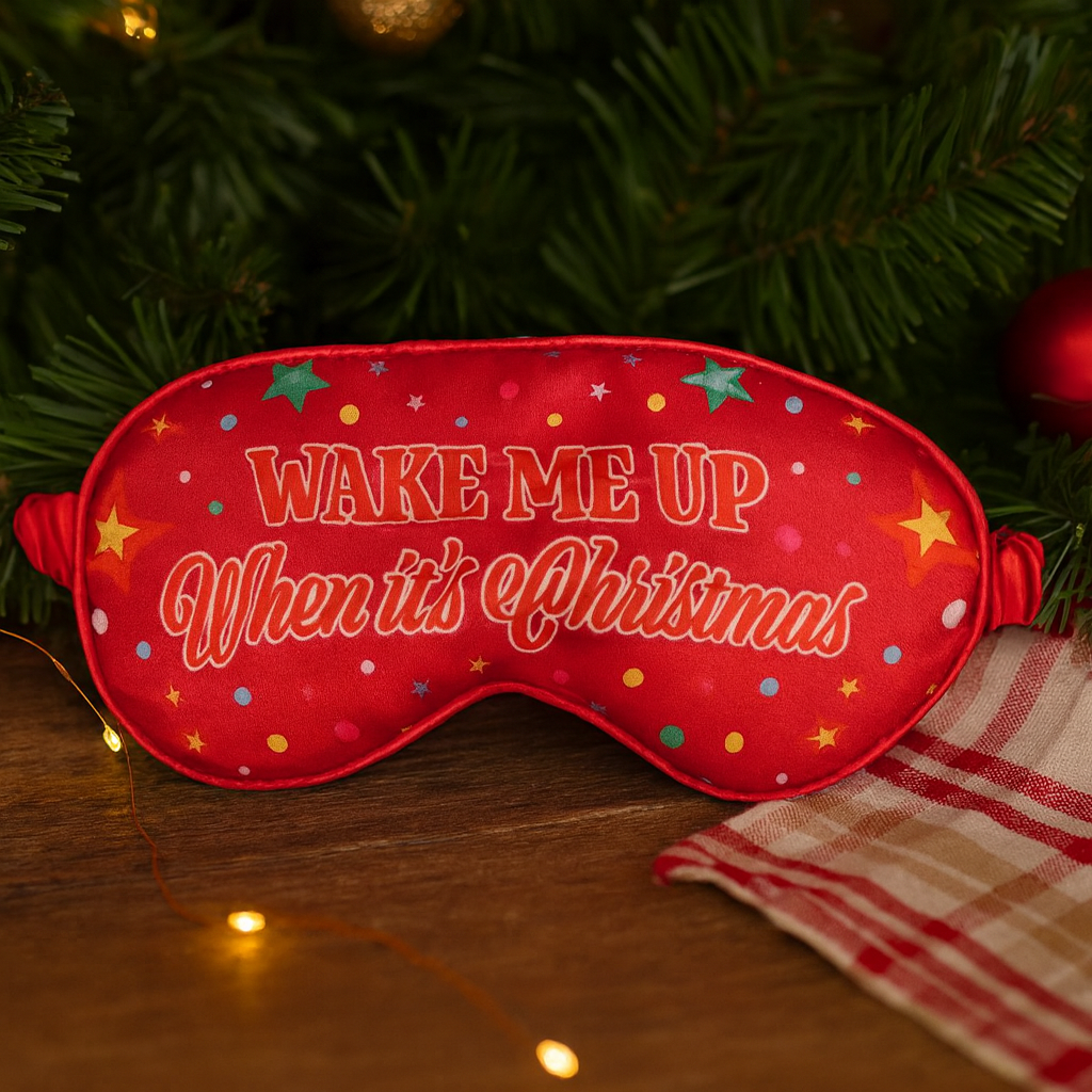 Red sleep mask with 'Wake Me Up When It's Christmas' text on a Christmas tree background