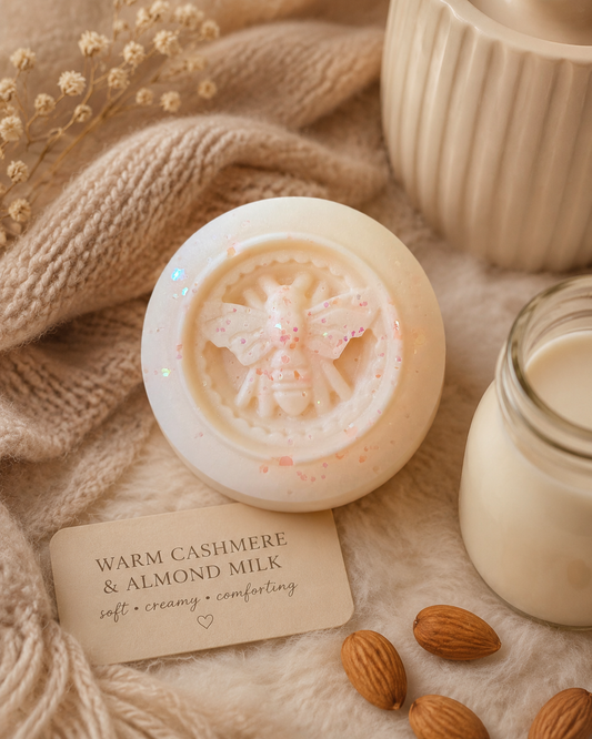 A round wax melt with a lid embossed with a bee, labeled 'Warm Cashmere & Almond Milk,' styled on a soft cream surface with a knitted beige scarf, dried flowers, almonds, and a glass jar of cream.